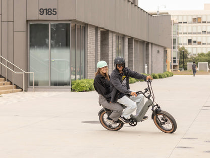 Two riders on a grey Troxus Lynx 2-Up electric bike with passenger seat and step-thru frame riding through an urban plaza