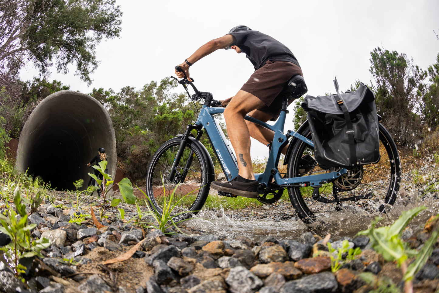 Rider on a Troxus Trax SUV electric bike in Cadet Blue crossing a creek with cargo pannier, showcasing comfort and all-terrain performance